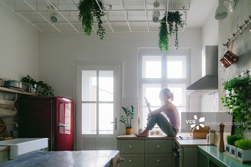 年轻女子坐在厨房台面上，用手机查看新闻。图片素材