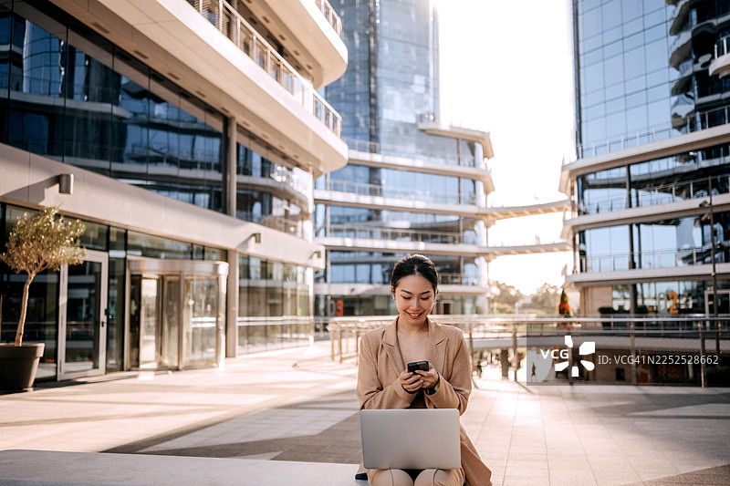 一位衣着光鲜的微笑女商人，坐在户外，膝盖上放着笔记本电脑，正在用智能手机发短信。图片素材