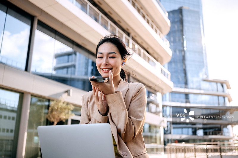 一位衣着光鲜的微笑女商人坐在户外，膝盖上放着笔记本电脑，正在用智能手机交谈图片素材