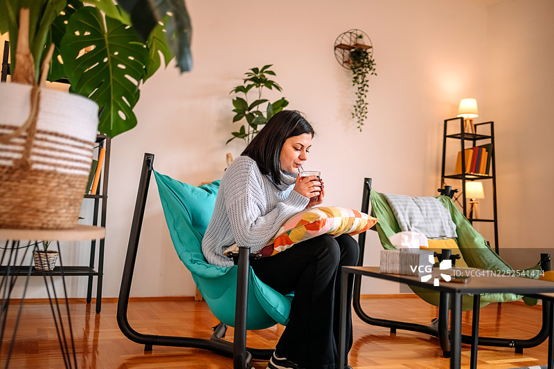 一位女士在家里的客厅检查健康状况，手中捧着茶杯取暖。图片素材