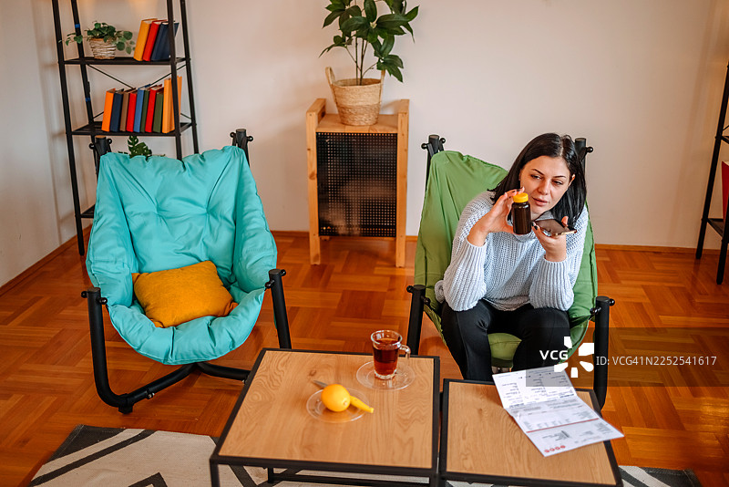 一名女子在家中的客厅里查看她的健康状况，一边看着药瓶，一边用手机寻求建议。图片素材