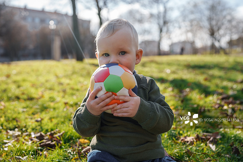 阳光明媚的日子里，一个年幼的孩子坐在公园的草地上，手里拿着一个五彩缤纷的球。图片素材