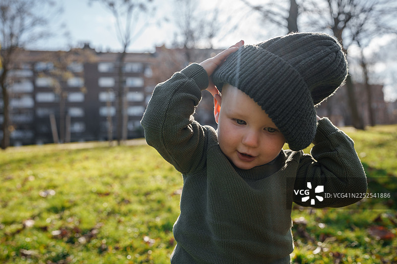 一个晴朗的日子里，在长满青草的城市公园里，一个小男孩开心地调整着一顶大大的针织帽子。图片素材