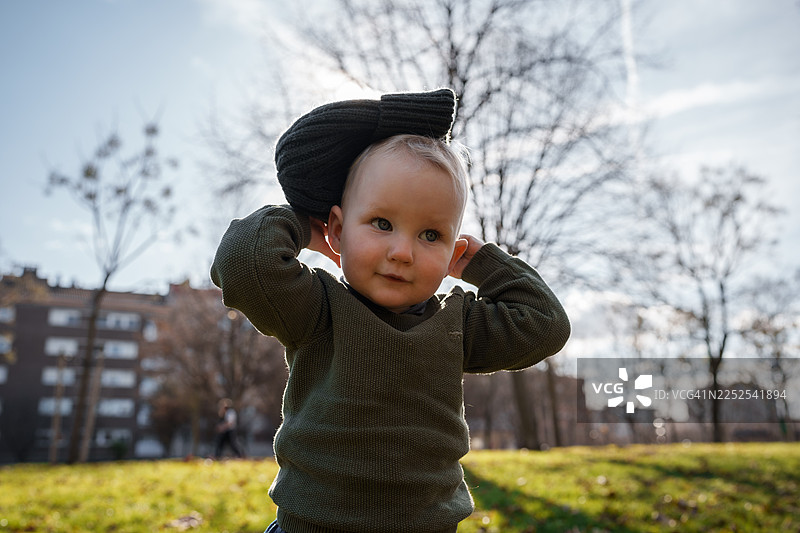 一个快乐的小孩在阳光明媚的公园里玩耍，周围是秋天的树木，他正举着一顶针织帽。图片素材