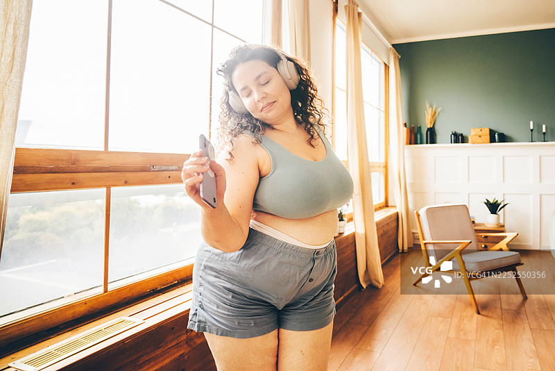 一位身材丰满的女性身穿蓝色家居服，戴着耳机，在阳光明媚的客厅里快乐地跳舞。图片素材