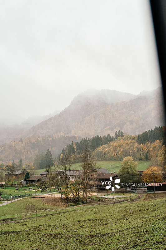 巴伐利亚阿尔卑斯山，一个乡村高山村庄，有农舍和绿色的草地，四周环绕着森林覆盖的山丘，在阴天的秋日里，部分笼罩在薄雾和低云中。图片素材