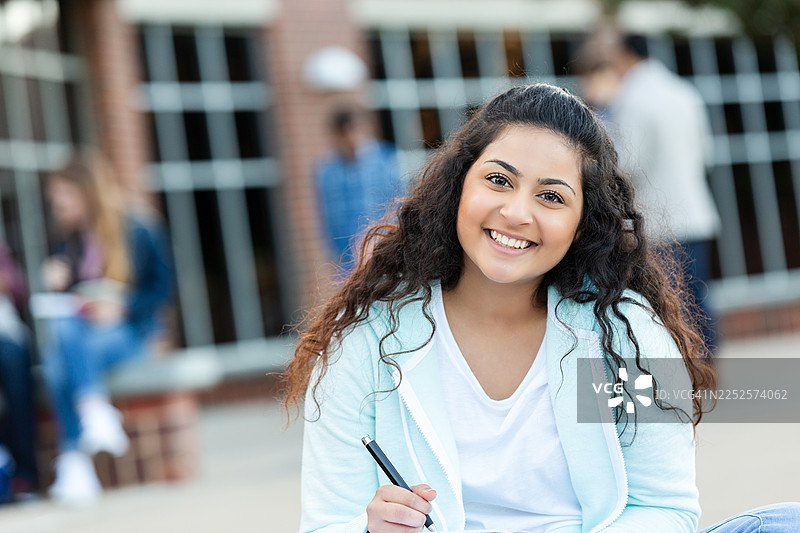 年轻的拉丁裔女学生在教学楼外微笑着用笔写作图片素材