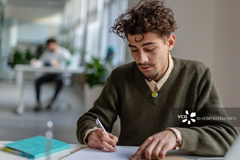 年轻人在白天的现代办公室环境中，在纸上写作，同事在工作图片素材