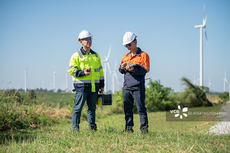 身着安全头盔和背心的工程师们在风电场工地检查数字平板电脑并使用对讲机沟通，风力涡轮机代表着可再生能源和可持续能源发电。图片素材