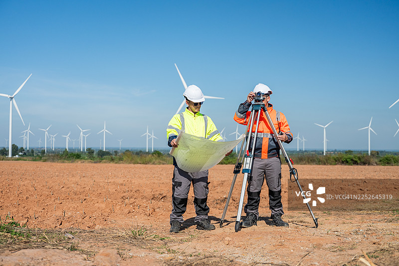 工程师在可再生能源现场的风力涡轮机旁工作，检查蓝图和安装在三脚架上的测量仪器，确保精确的土地测量和环保的工业实践。图片素材