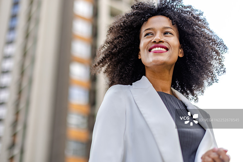 年轻的黑人女性在城市环境中面带微笑，身穿浅色夹克，站在高楼大厦前。图片素材