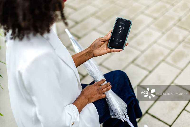 一位年轻的黑人女性在阴天户外，身边放着雨伞，她正坐在那里用手机查看天气。图片素材