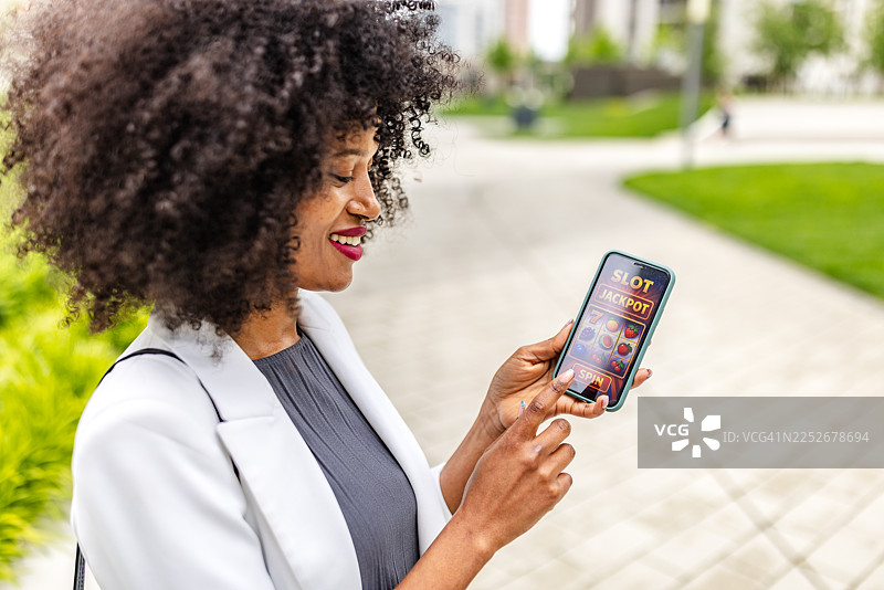 年轻女子白天在城市公园户外玩智能手机上的老虎机游戏时面带微笑图片素材