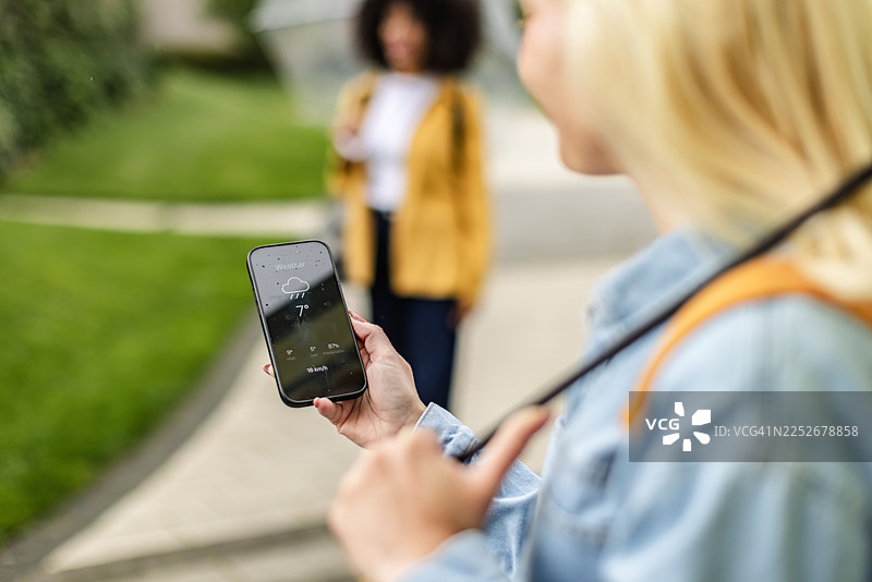 年轻的黑人女性和年轻的白化病女性在公园里享受户外时光，并用智能手机查看天气图片素材