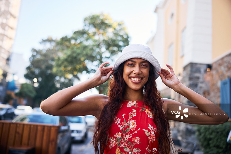 一位年轻女子穿着碎花连衣裙，在阳光明媚的城市街道上，边微笑边调整她的帽子。图片素材