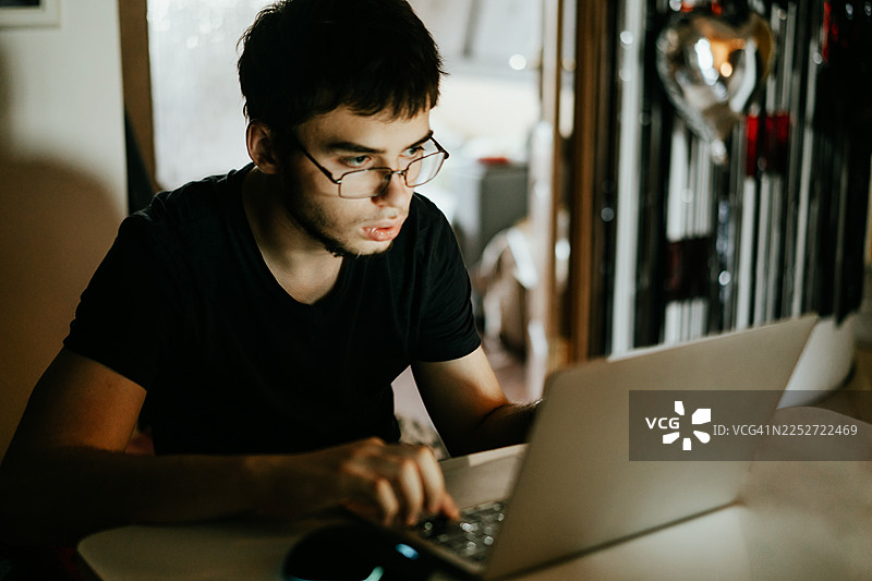 年轻男子在家中光线昏暗的房间里，专注地使用笔记本电脑学习工作图片素材