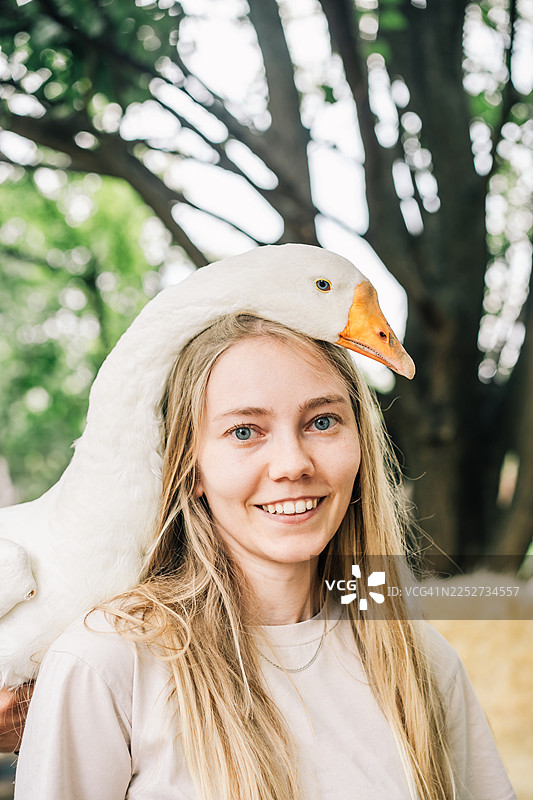 一位年轻女子在户外的肖像，一只白鹅轻轻地停在她的头上，在自然日光下营造出一种俏皮而温柔的农场生活瞬间。图片素材