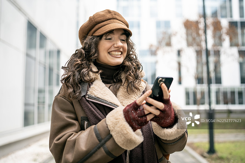 穿着舒适的棕色羊羔毛外套的女性在城市户外使用智能手机，面带微笑图片素材