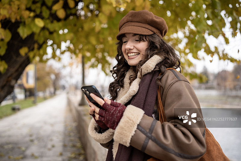 一位穿着温暖秋季外套和帽子的女士，在河边风景优美的公园里使用智能手机，脸上带着笑容图片素材