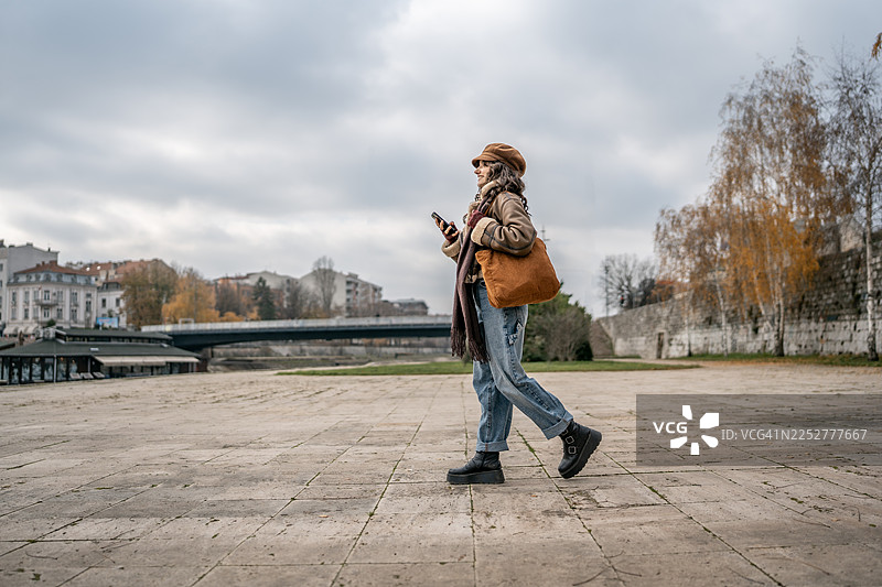 身穿牛仔裤、靴子，手提棕色包包的时尚女子在秋季的城市中行走，手中拿着手机图片素材