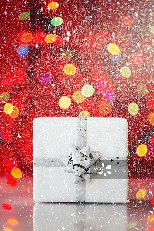 放在反光面上的白色礼品盒，带有银色蝴蝶结，背景为红色，有失焦的灯饰。新年即将来临的概念。正面视角，特写。图片素材