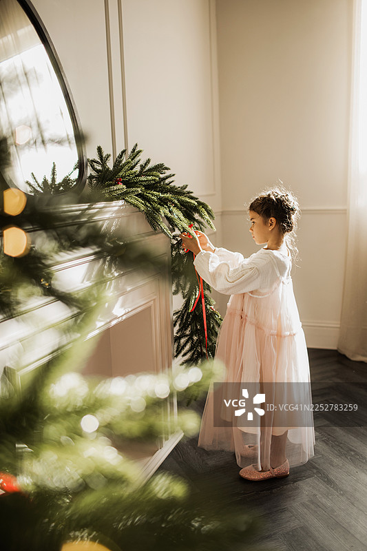 穿着粉色连衣裙的小女孩在舒适的家中装饰壁炉上的圣诞花环图片素材