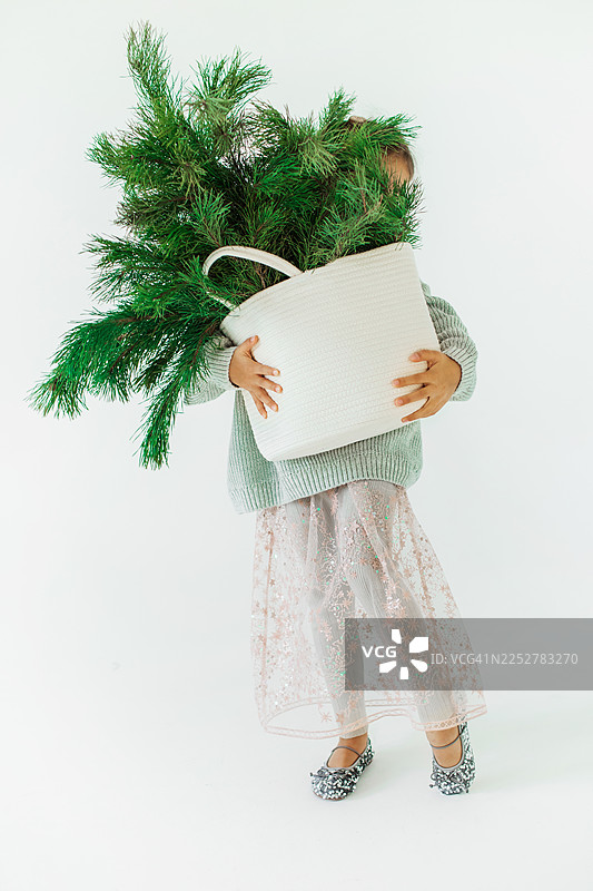 孩子抱着装有圣诞树的白色篮子，身穿舒适的冬季服装，温馨的节日时刻图片素材