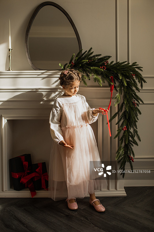 身穿白裙的小女孩站在挂着红丝带和礼物的节日花环拱门旁图片素材