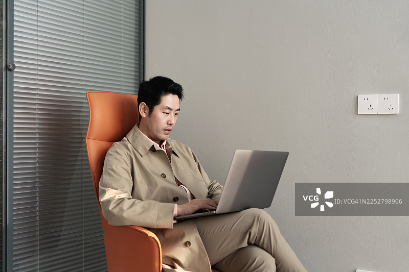 年轻的亚裔商人坐在办公室的橙色沙发上使用笔记本电脑。图片素材