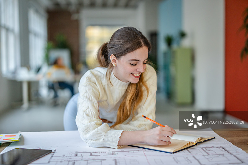 年轻女子在现代办公室工作，同事们在忙于各种事务，她在一本笔记本上书写图片素材