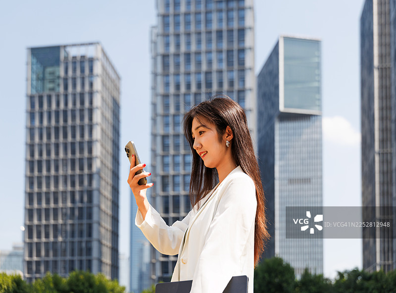 一位成功的年轻亚洲女商人在城市中央商务区，身着现代职业装，手持智能手机和笔记本电脑，穿梭于高耸的现代企业大楼之间，体现了职场女性的领导力以及随时随地处理工作的商务精神。图片素材