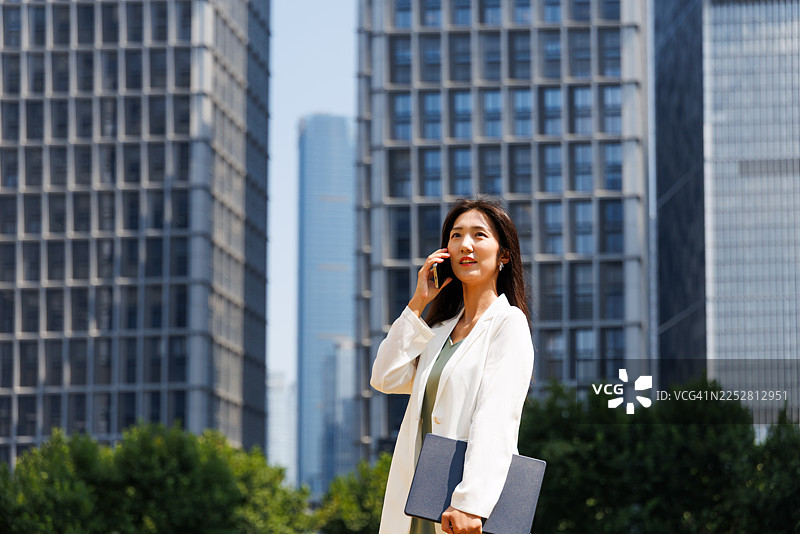 一位成功、现代的年轻亚洲女商人，手持智能手机和笔记本电脑，在现代企业建筑林立的城市中央商务区通勤上班。女性领导力。移动办公。图片素材