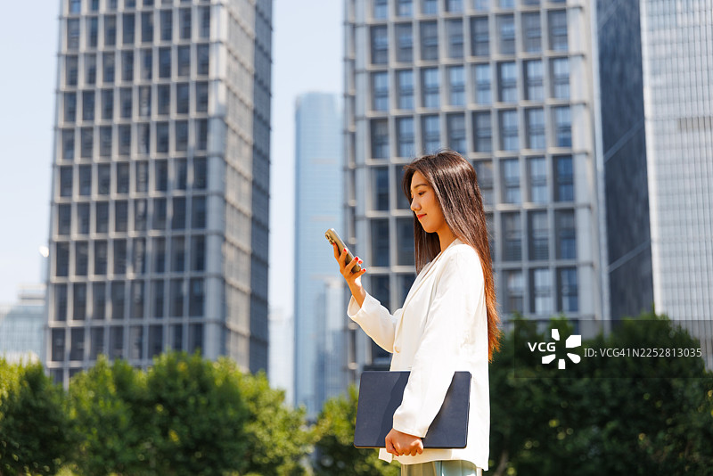 一位成功的现代亚洲年轻女商人，手持智能手机和笔记本电脑，在城市中现代化的企业建筑背景下，前往中央商务区通勤。女性领导力，移动办公。图片素材