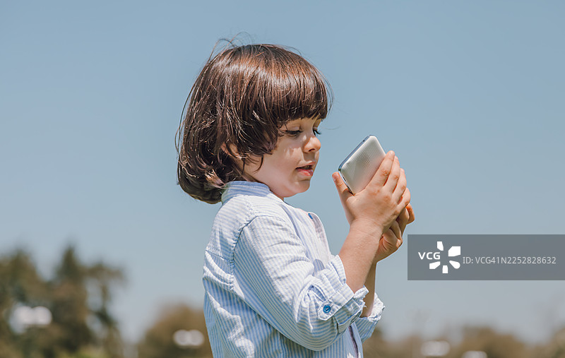 一个阿拉伯小男孩在阿联酋迪拜的绿色公园里，一边使用智能手机一边打电话。图片素材
