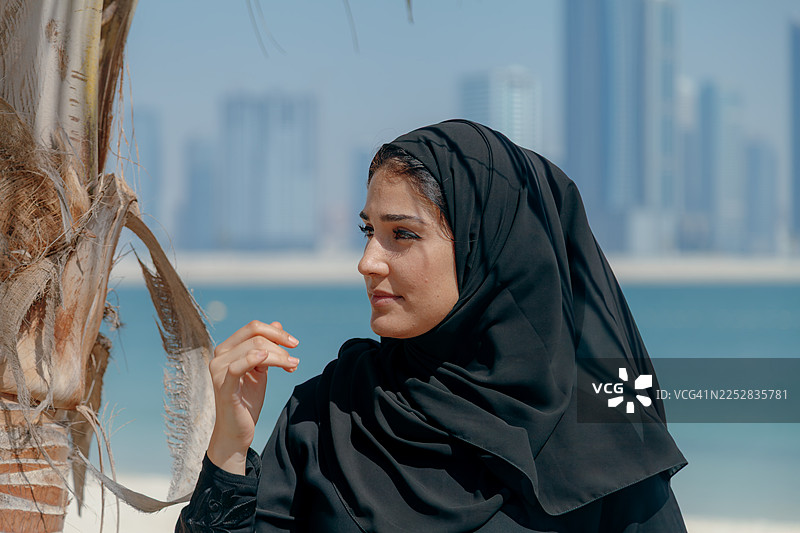 迪拜阿联酋，阳光沙滩棕榈树旁，身着黑色阿巴亚的年轻中东女性面带微笑，背景是现代城市天际线图片素材