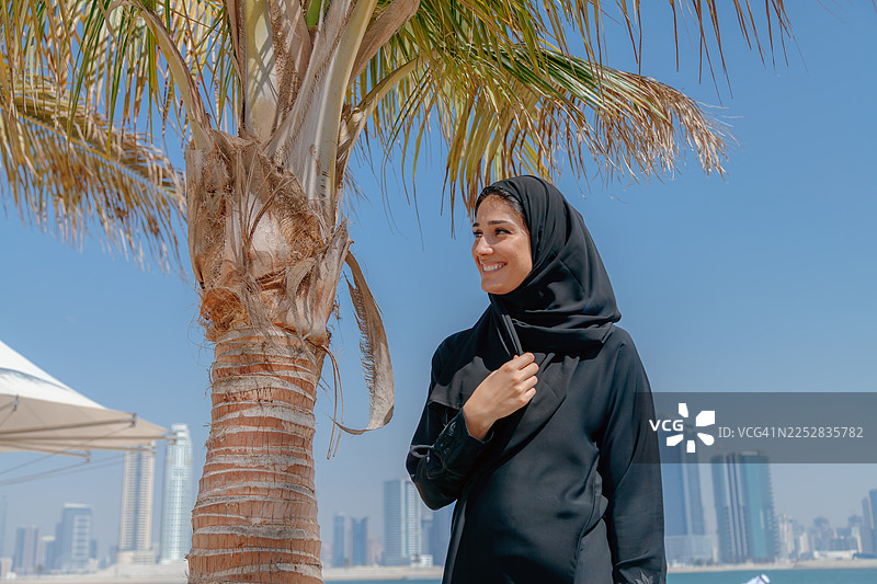 阳光明媚的海滩上，一位身穿黑色阿巴亚的年轻中东女子站在棕榈树旁，背景是迪拜的现代城市天际线。图片素材