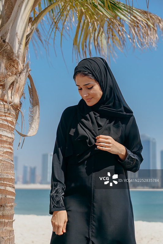迪拜阿联酋，阳光明媚的海滩边，一位身穿黑色阿巴亚的年轻中东女性站在棕榈树旁，背景是现代化的城市天际线，她面带微笑。图片素材