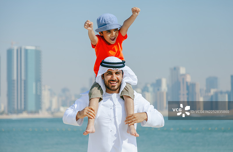 一位微笑的阿拉伯父亲，在背景为迪拜天际线的沙滩上玩耍时，将年幼的儿子扛在肩上。图片素材