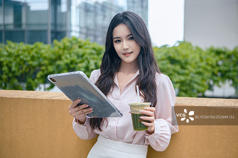 在现代摩天大楼露台上，一位专注的亚洲女商人一边使用平板电脑工作，一边拿着一杯绿色果汁。自信的职业女性在户外使用iPad，背景是城市景观，体现专注和高效的概念。图片素材