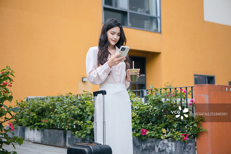 年轻的亚洲女性在城市花园露台上，带着行李使用智能手机，享受现代都市生活、旅行、休闲和户外数字连接。图片素材