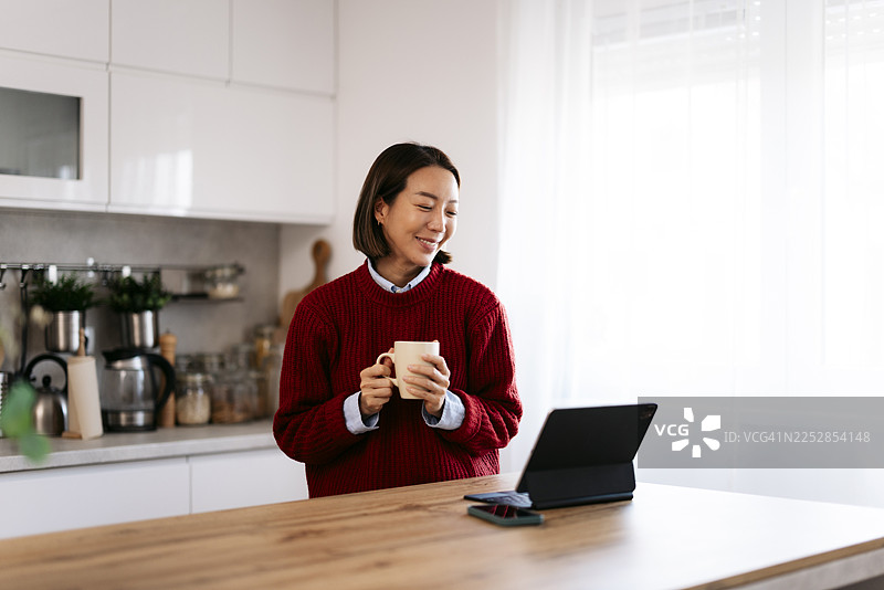 亚洲女性在家里的厨房里，一边享用咖啡，一边远程工作图片素材
