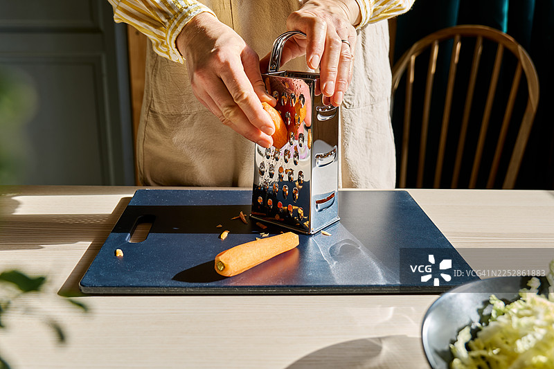 一位看不清面容的女士正在砧板上用擦丝器擦新鲜胡萝卜。正在烹饪健康食品。图片素材