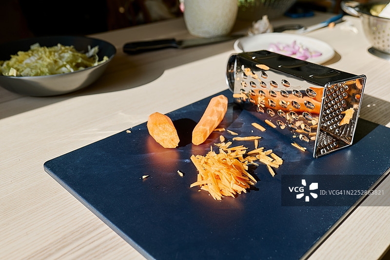 桌上放着切菜板上的胡萝卜和擦丝器，碗里装着切好的北京白菜，盘子里装着切好的洋葱。烹饪健康食品。图片素材