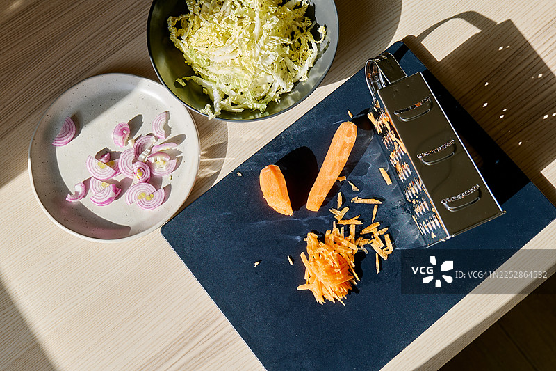 案板上胡萝卜靠近刨丝器，碗里装着切好的白菜，盘子里装着切好的洋葱，都在桌子上。烹饪健康食品。图片素材