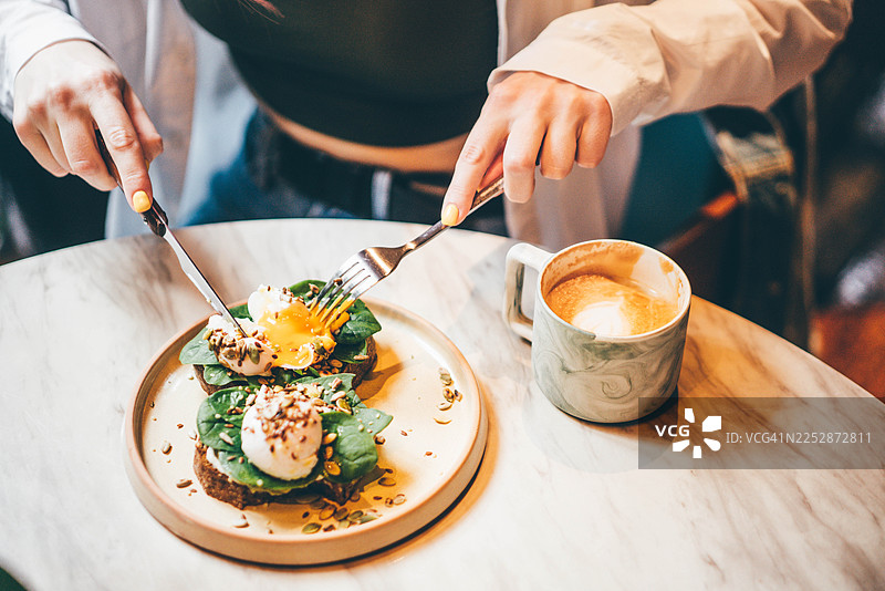 一位女士在咖啡馆的大理石桌上，用刀叉享用着牛油果吐司和水波蛋的特写镜头。图片素材