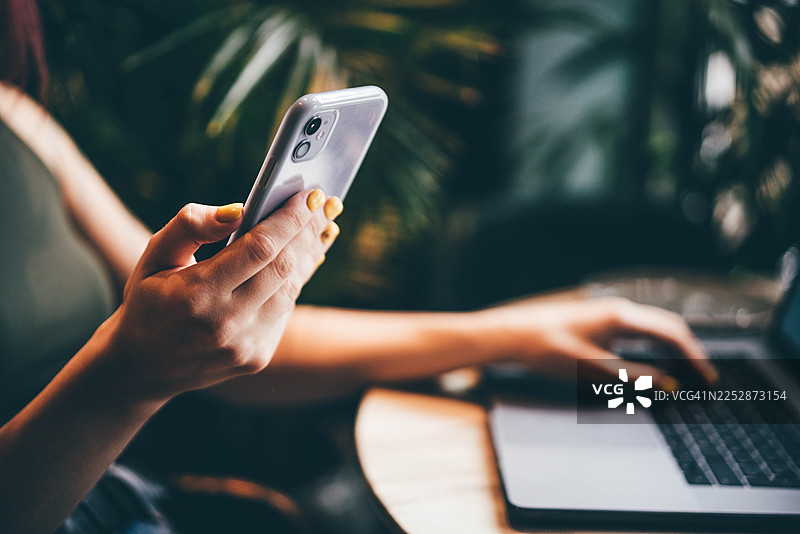 特写镜头，一位女士在咖啡馆或家庭办公室，一边使用笔记本电脑一边用手机。这张图片代表了数字连接、远程工作和现代忙碌生活方式的概念。图片素材