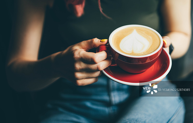 一位女士手捧着装有精美心形拉花拿铁的红色杯子的特写。图片素材