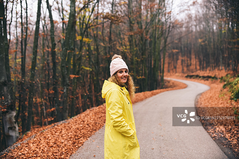 穿着亮黄色雨衣的女子走在落满树叶的秋季森林小径上图片素材