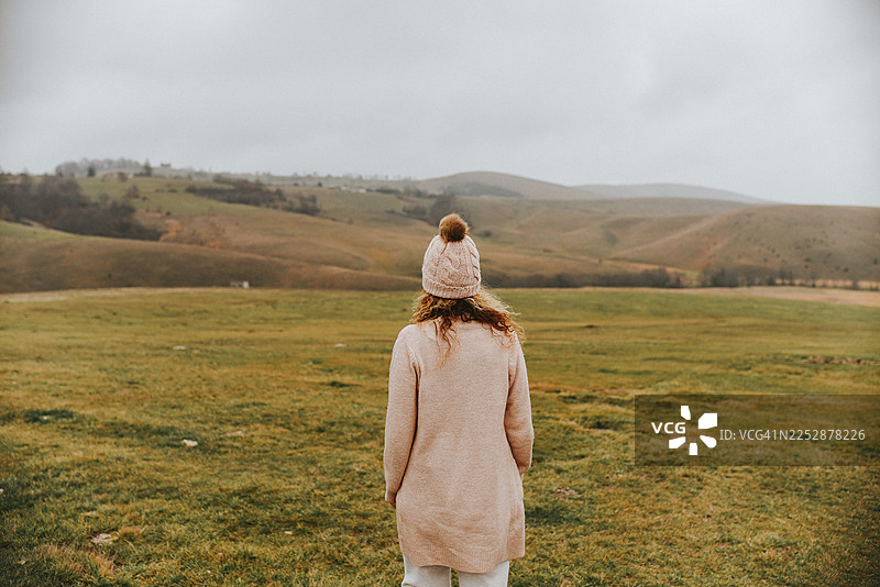 身穿米色外套和针织帽的女子独自站在多云天空下的开阔田野中图片素材