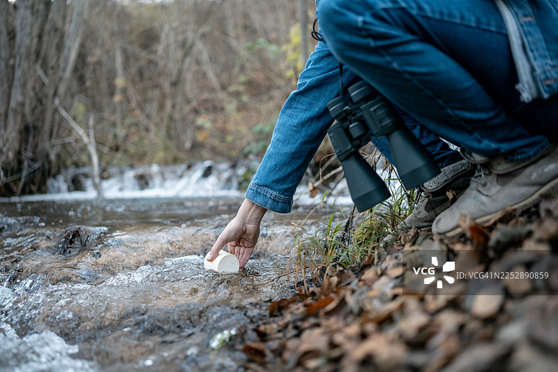 秋季自然风光中，一个人跪在溪流旁，手持双筒望远镜，采集水样。图片素材